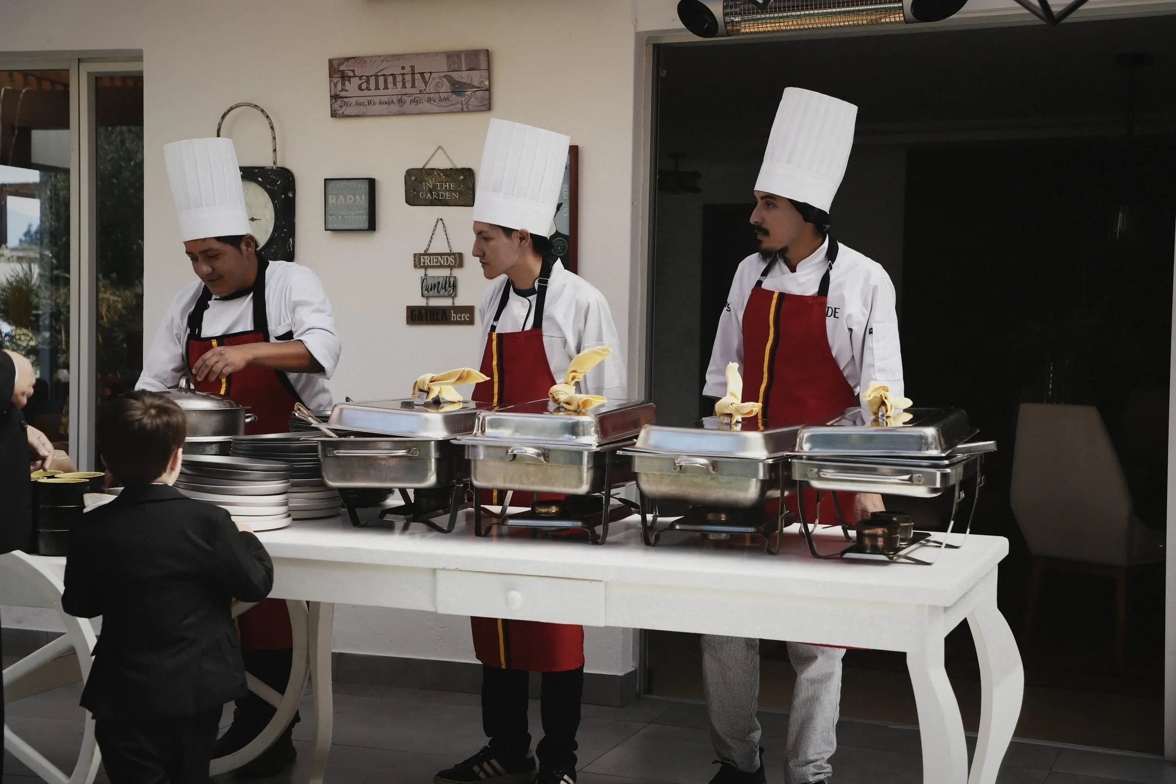 Three cooks in red aprons serving from a line of chafing dishes.